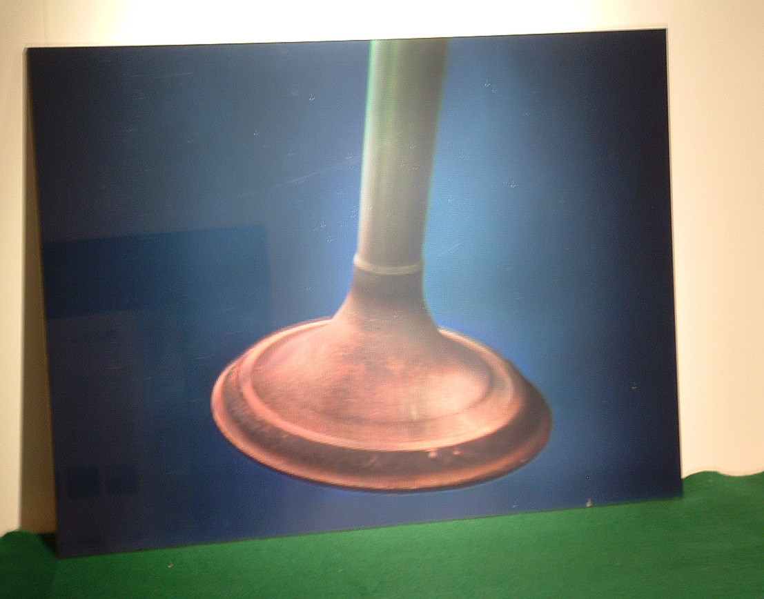 Physical lenticular hologram of a clean engine valve, mounted on a stand and photographed against a soft studio backdrop.